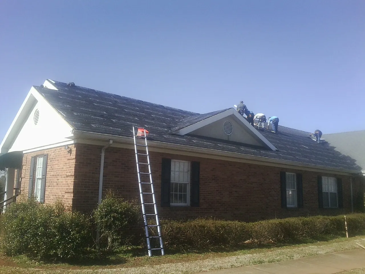 Crew tearing shingles off a brick ranch roof with a ladder set — Metal Roof Repair work by our crew in Mc Grann