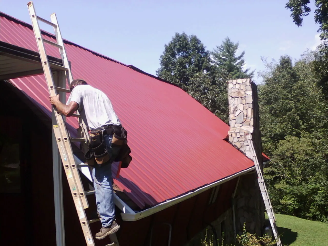 Roofer on a ladder beside a red metal roof and stone chimney — Soffit & Fascia Repair by Roofing Brothers in Mc Grann